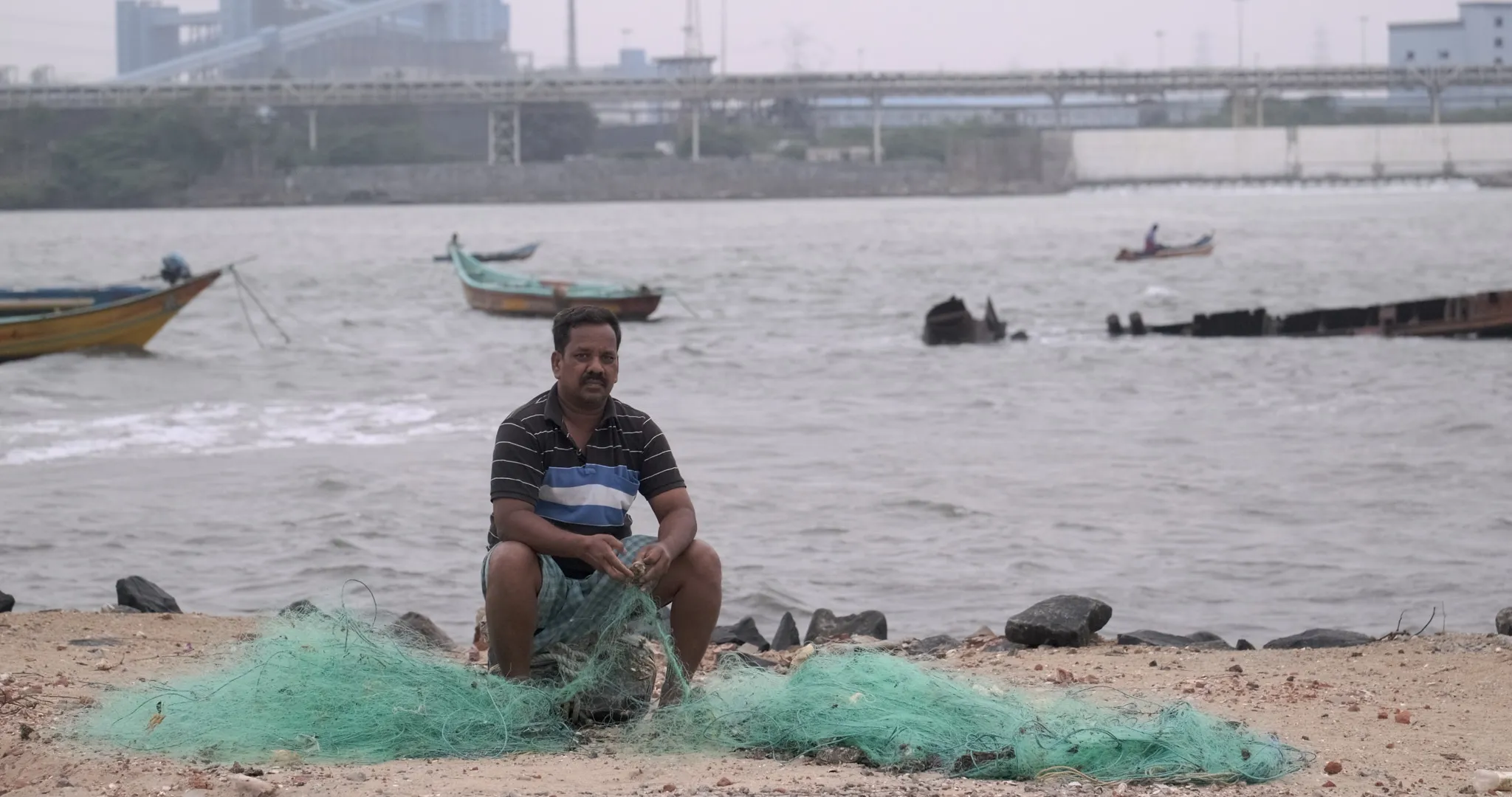 Rajan, a fisherman from Nettukuppam village in Ennore, mends his fishing net with a coal-based power plant (North Chennai Thermal Power Station) in the background. He says that fish catch has declined in the neighbouring villages due to hot water and effluent discharge from the power plant.